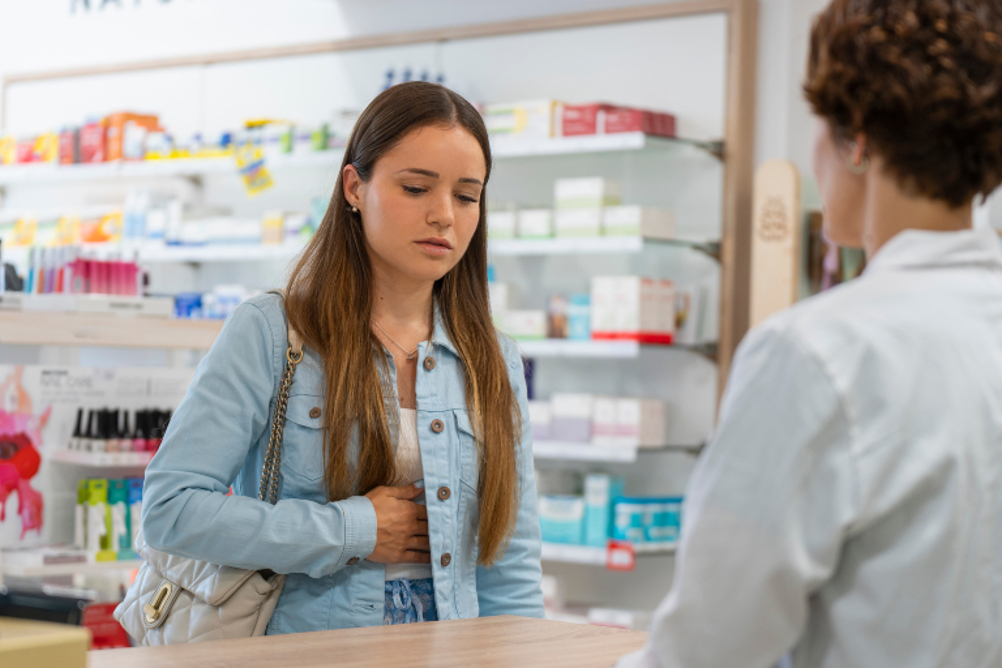 Young Woman Holding Her Stomach To Illustrate A Stomach Ache Speaking To A Pharmacist At A Pharmacy