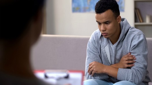 A young man looking downcast in a consulting room while being looked on by a professional holding a clipboard